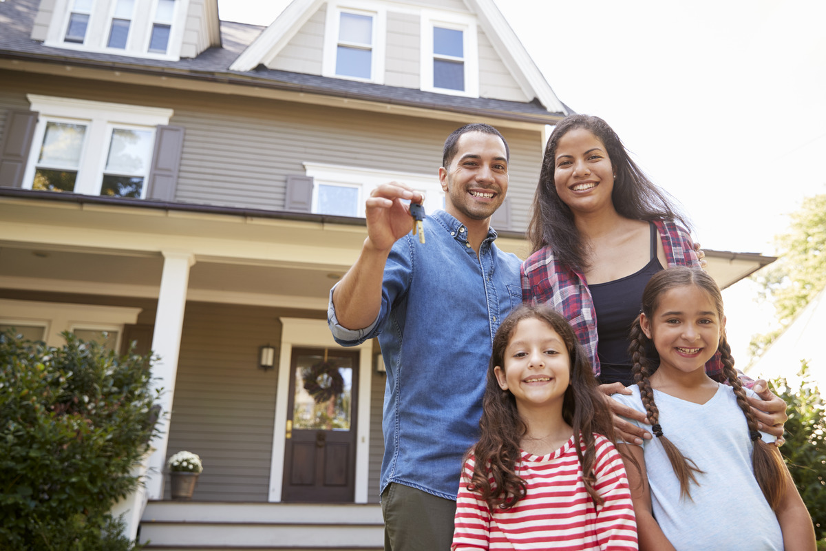 Husband wife and two children holding new keys to their new home behind them with a dark colored door with wreath hanging.